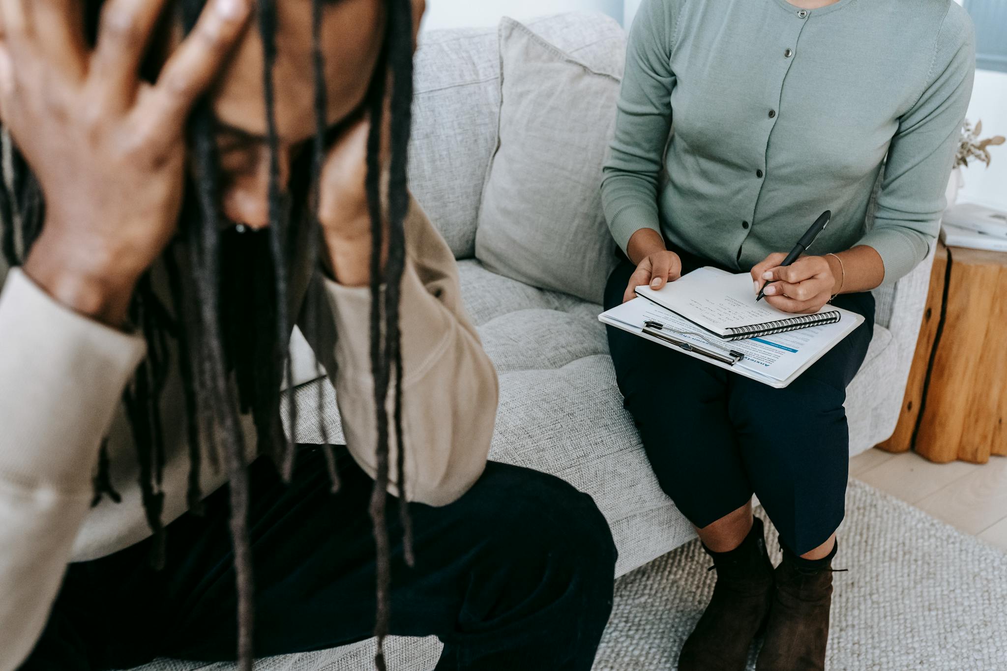 Crop concerned African American male sitting on sofa with female psychologist and holding head in hands in dismay during psychotherapy session